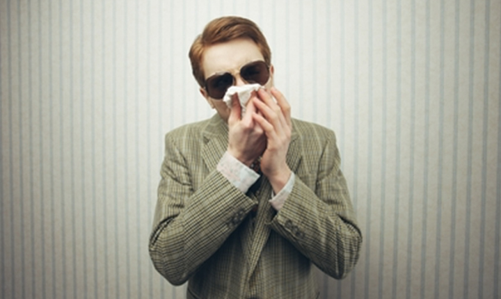 Man wearing sunglasses and a checkered suit, holding a tissue to his nose.