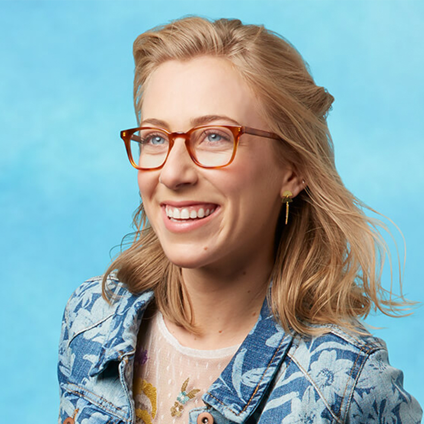 Woman wearing rectangular, amber-colored eyeglasses, a floral denim jacket, and gold drop earrings.