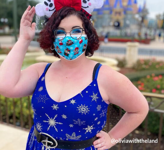 Person wearing a blue dress with white snowflakes, a festive mask, glasses, and Minnie Mouse ears. @oliviawiththeland