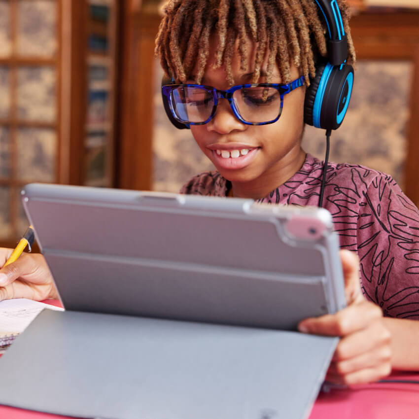 Child using a tablet with a gray case, wearing blue-rimmed glasses and black headphones with blue accents.