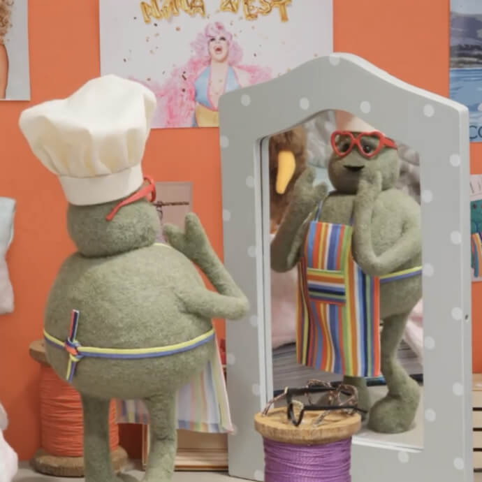 Green plush character wearing a chef’s hat and striped apron, admiring itself in a mirror with red glasses.