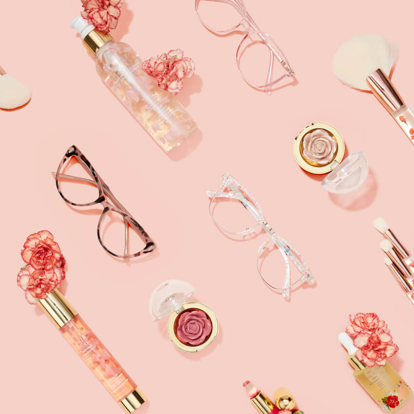 Assorted cosmetics and eyeglasses, including lipsticks, blush, brushes, and spray bottles on a pink background.