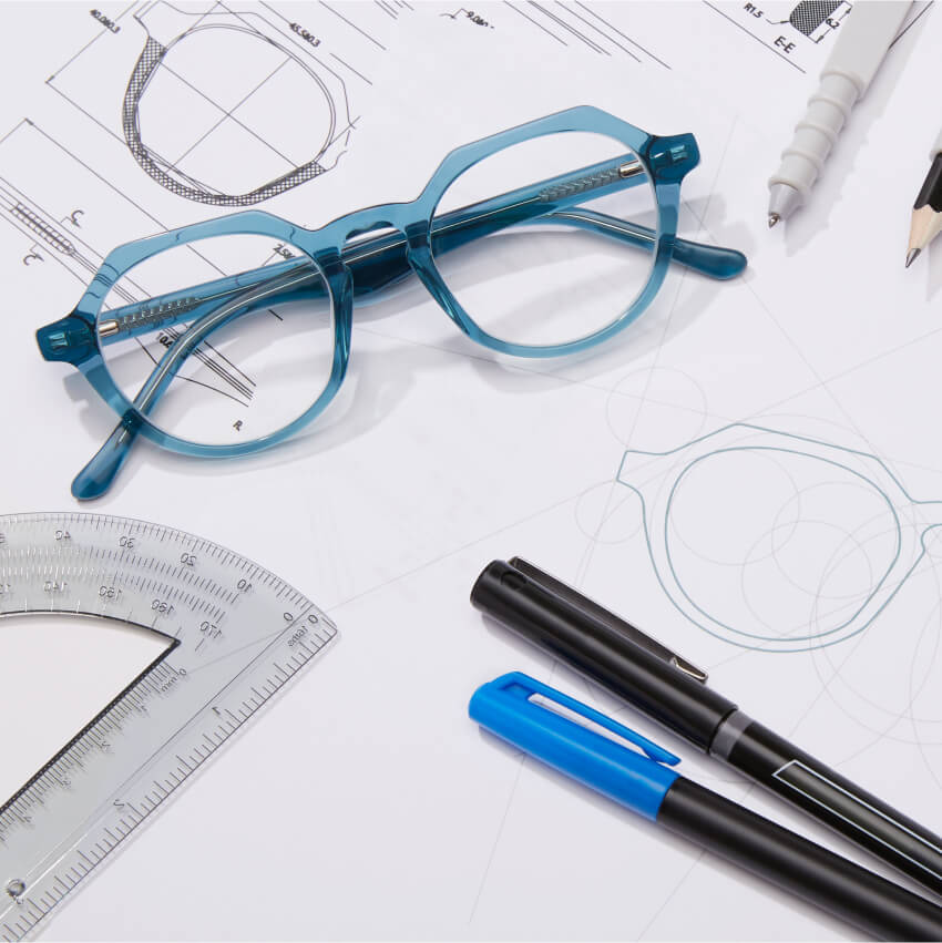 Blue eyeglass frame on technical drawing papers with a protractor, pens, and pencils.