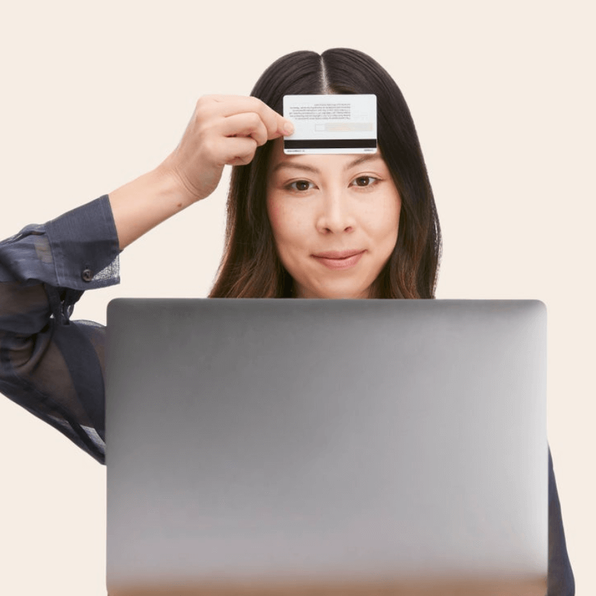 Woman holding a card above a laptop, showing card details for verification. Text on card is not readable.