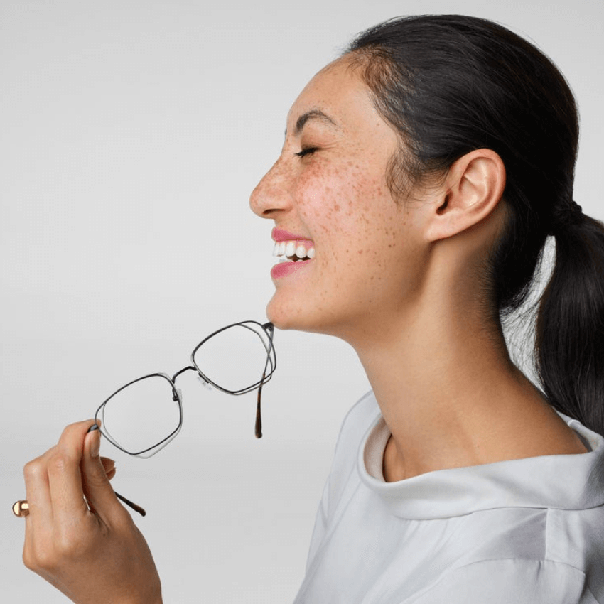 Woman holding black-framed eyeglasses, smiling with her eyes closed.