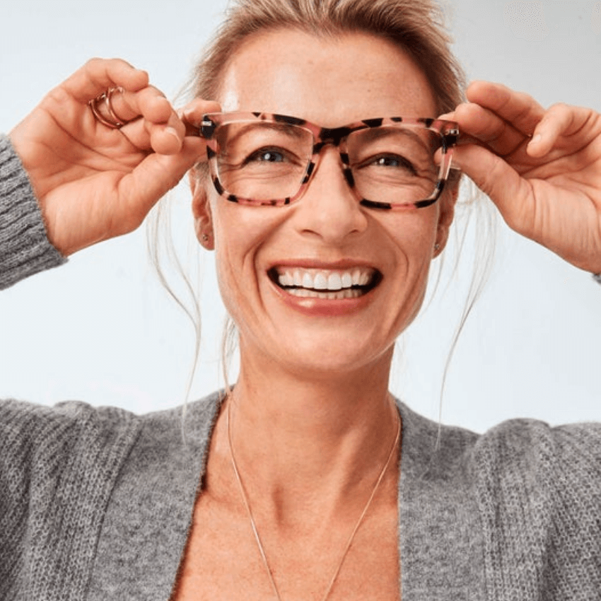 Woman smiling while holding tortoiseshell-patterned eyeglasses on her face.