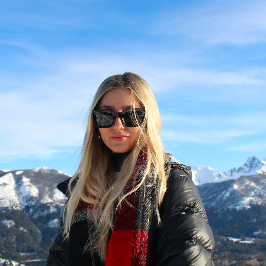 Woman wearing black sunglasses, a puffer jacket, and a red and black checkered scarf in a snowy mountain setting.