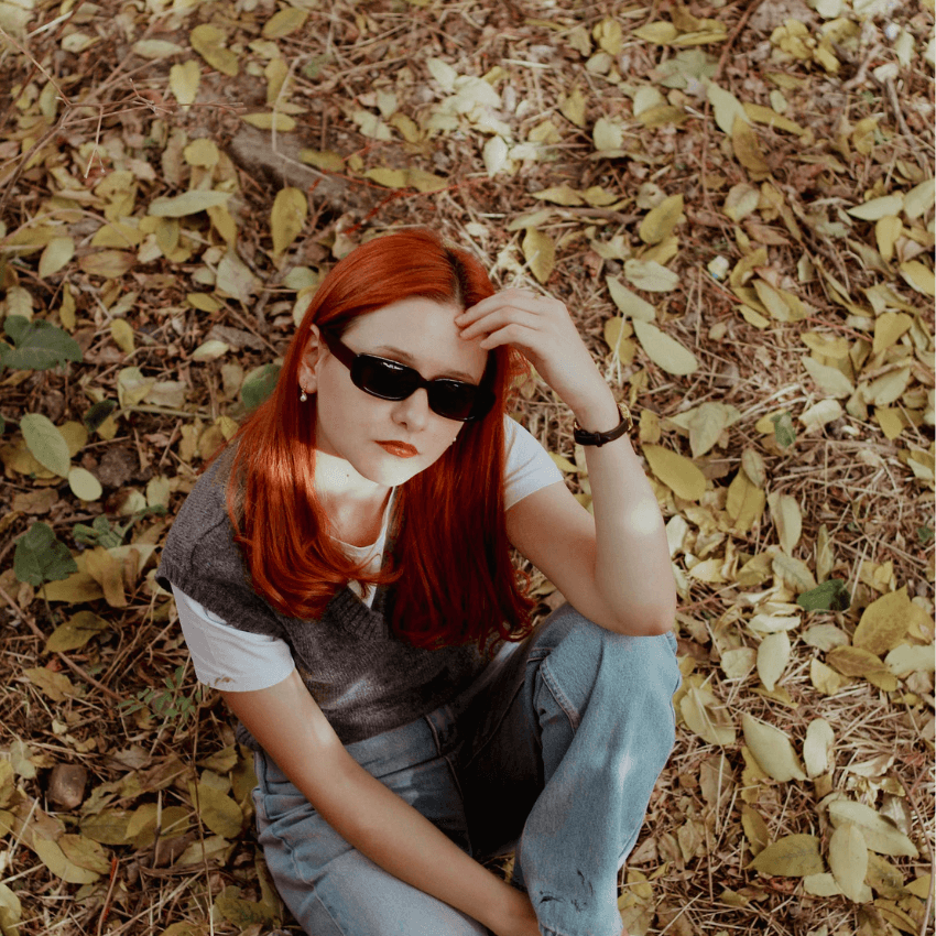 Person with red hair wearing black sunglasses, a gray vest over a white shirt, and blue jeans.