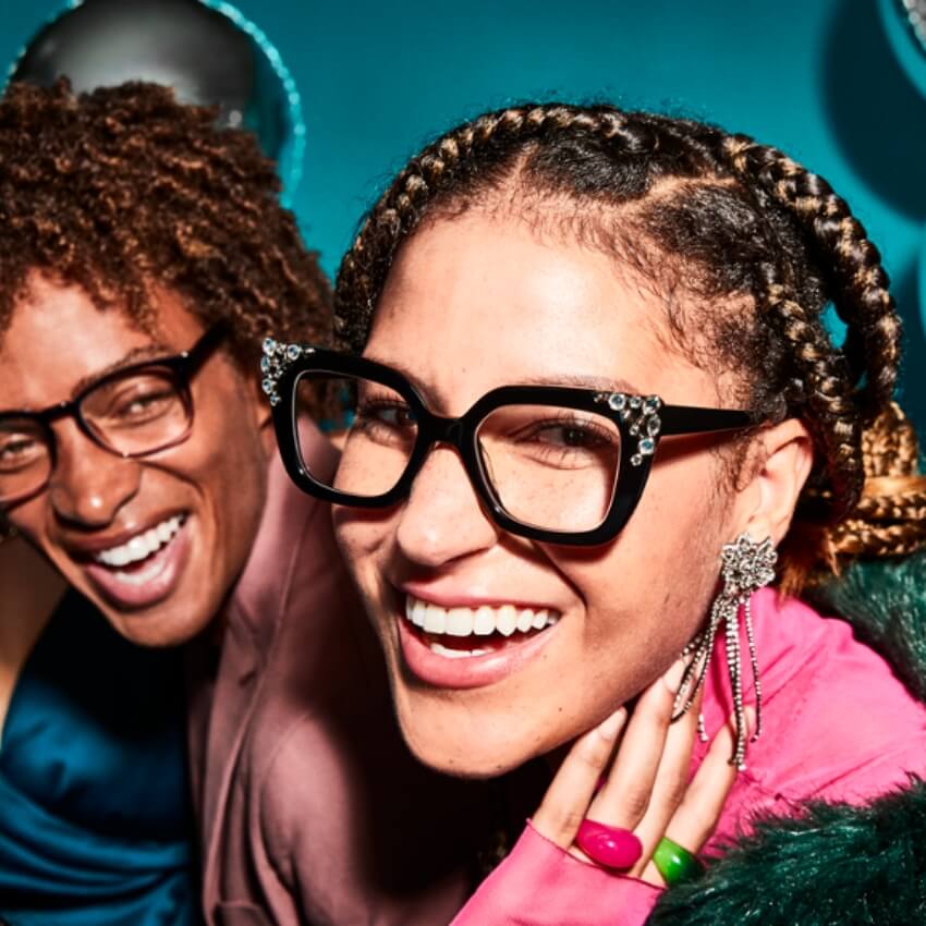 Smiling person wearing black rhinestone-studded glasses, pink top, large floral earring, and bright rings.