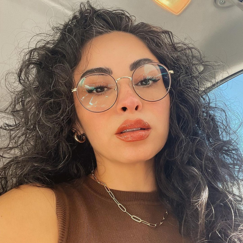 Woman with curly hair wearing round glasses, large hoop earrings, brown top, and gold chain necklace.