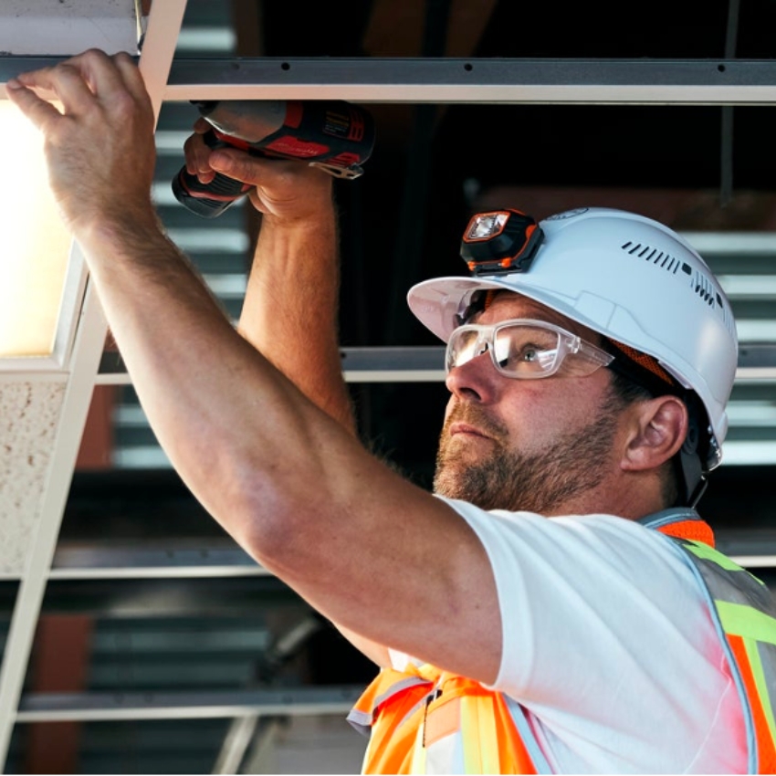Worker in hard hat and safety goggles using a drill to fix a light fixture on the ceiling.