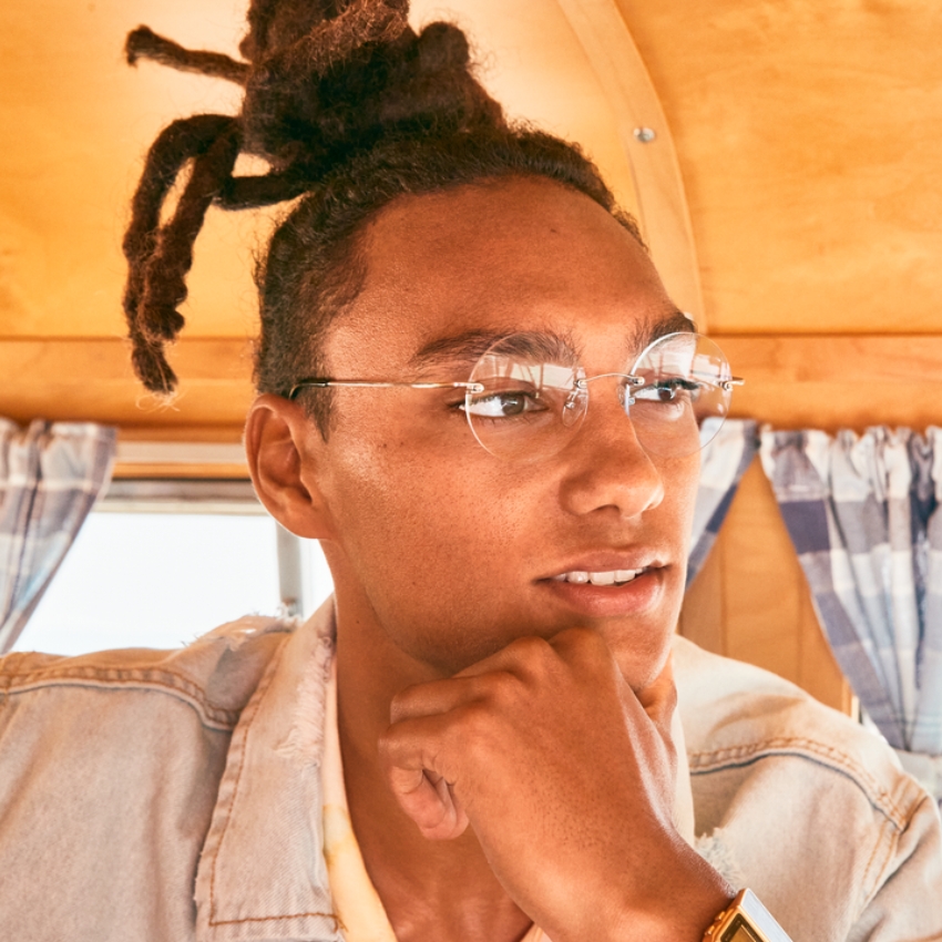Person wearing frameless glasses with thin metal arms and a gold wristwatch, resting chin on hand.