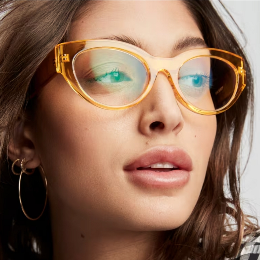 Woman wearing large, yellow translucent cat-eye glasses with light brown hair and gold hoop earrings.