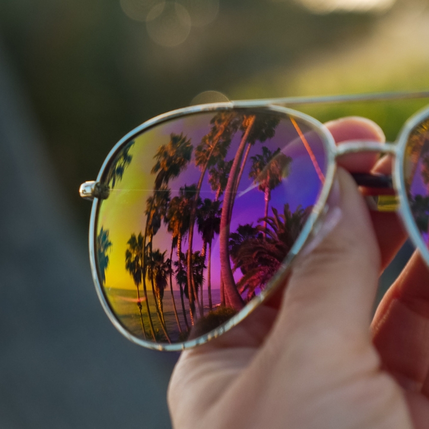 Reflective sunglasses with palm trees and colorful sunset scenery in the lenses held by a hand.