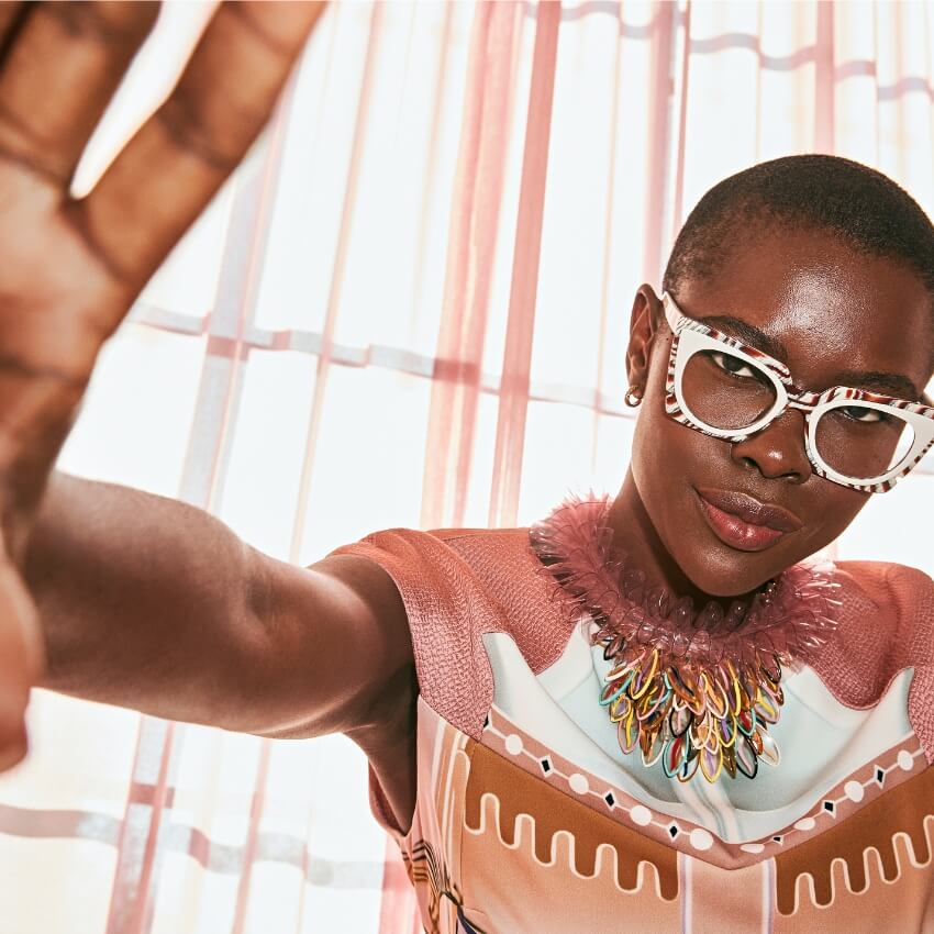 Model wearing large white patterned glasses, colorful statement necklace, and a patterned dress with pink tones.