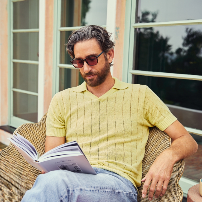 Man in a yellow polo shirt reading a book while wearing sunglasses.