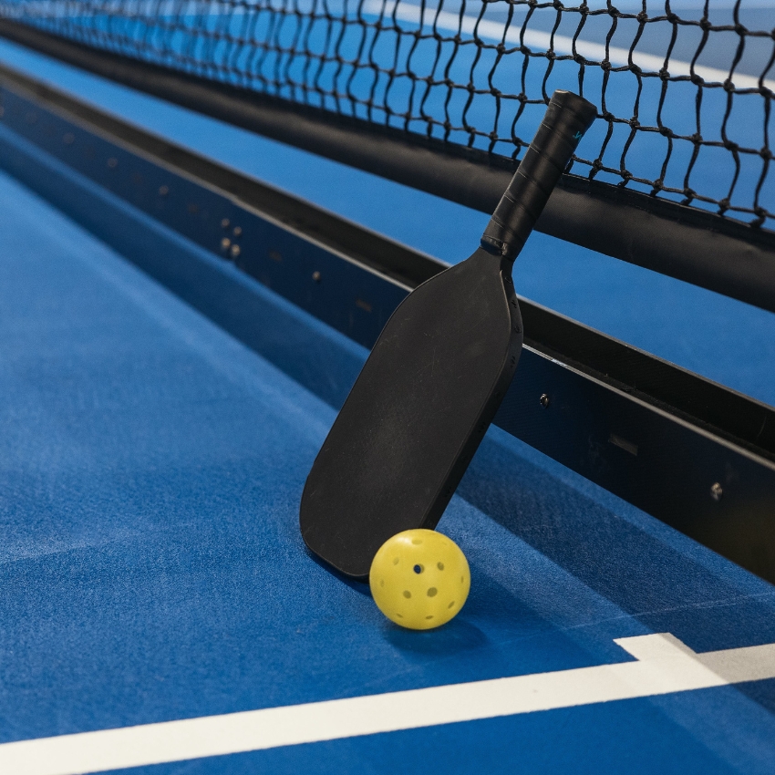 Black pickleball paddle leaning on a net with a yellow perforated ball on a blue court.
