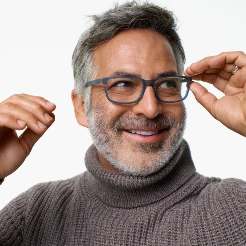 Man smiling while adjusting grey eyeglasses, wearing a grey turtleneck sweater. Plain white background.