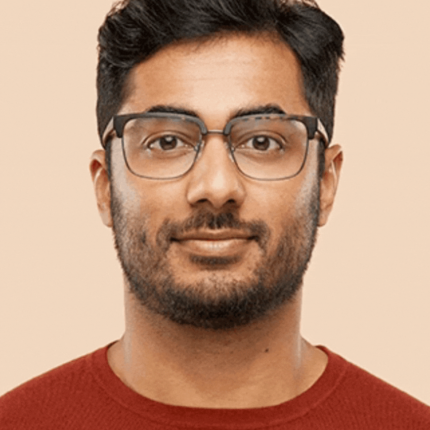 Man wearing black-rimmed eyeglasses and a red sweater against a plain background.