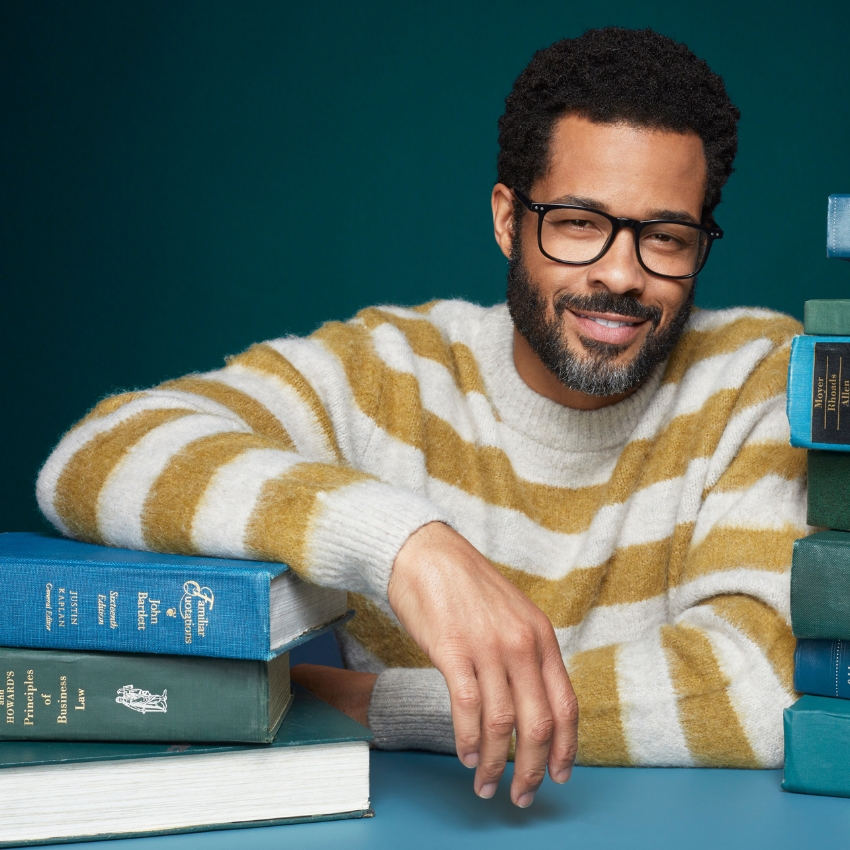 Man in striped sweater wearing glasses, leaning on a stack of books including "Jeff Koons Retrospective," "John Barth: A Body," and "Business Law."