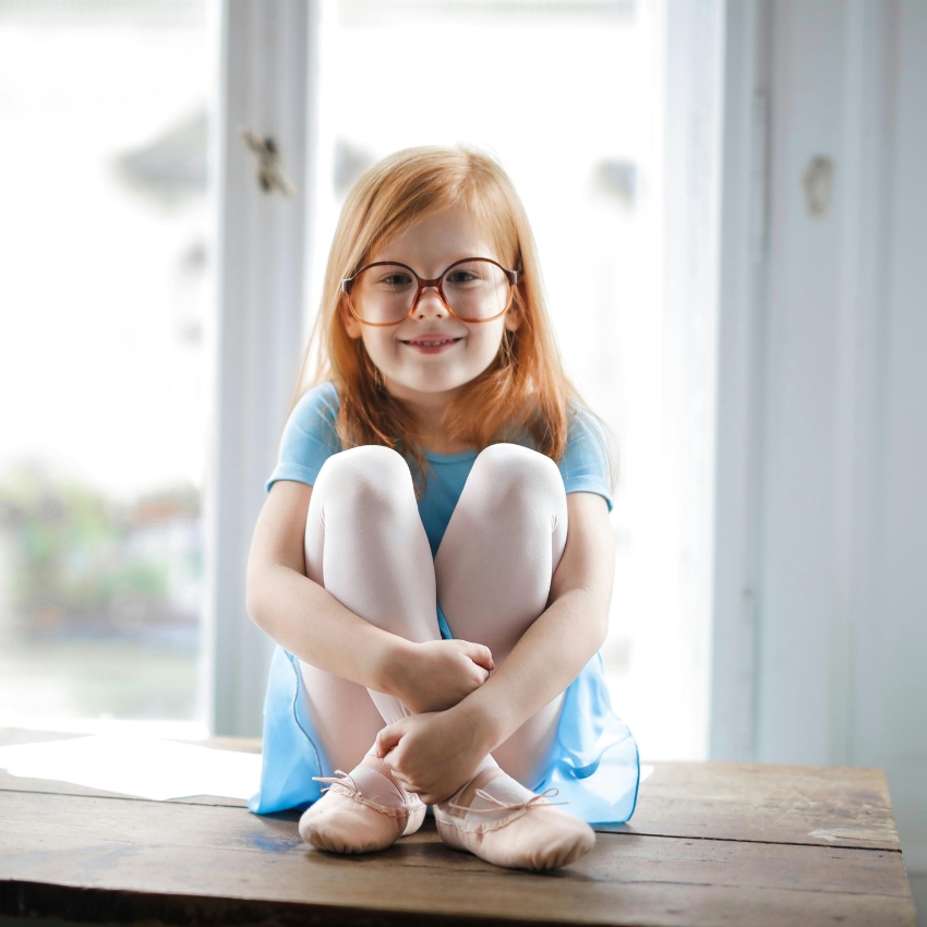 Child in glasses wearing a light blue dress, white tights, and pink ballet shoes while sitting.