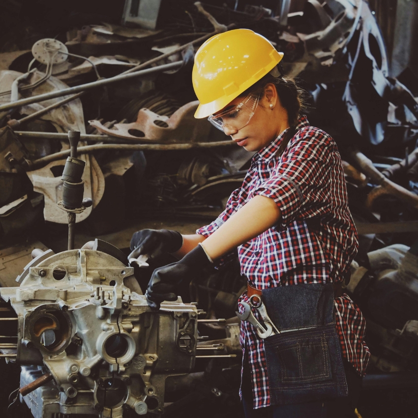 Worker in safety gear adjusting machinery, wearing yellow hard hat, safety glasses, and orange plaid shirt.