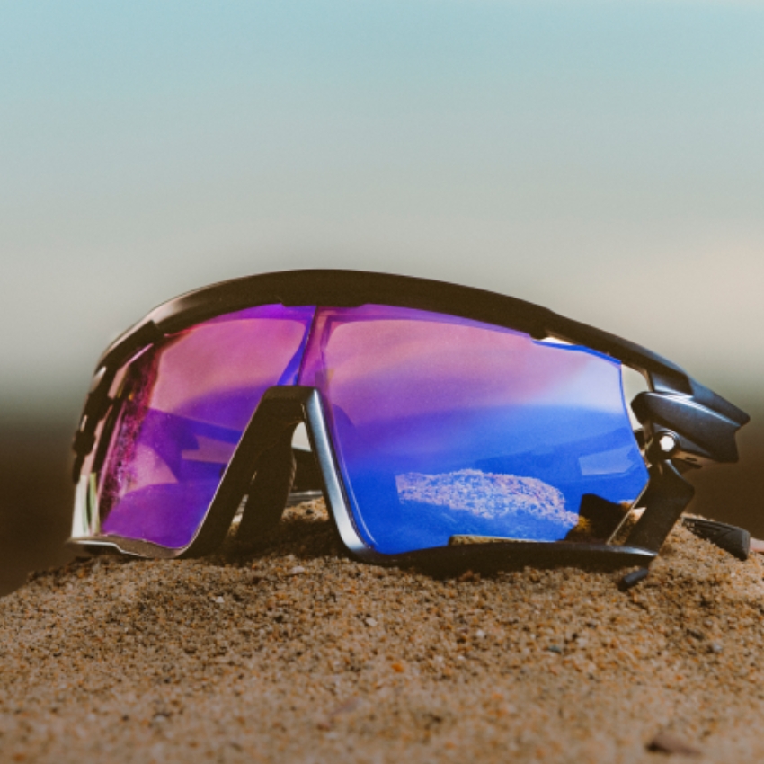 Sporty sunglasses with purple and blue mirrored lenses resting on a sandy surface.