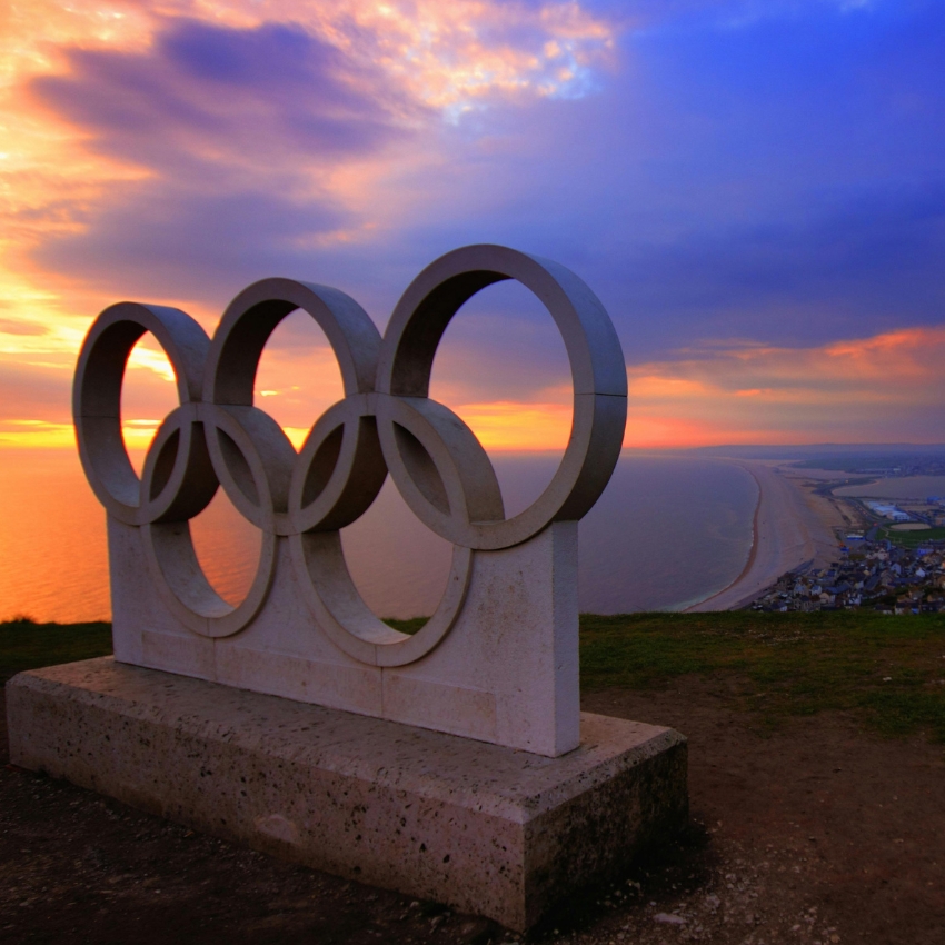 Olympic rings sculpture with coastal sunset background.