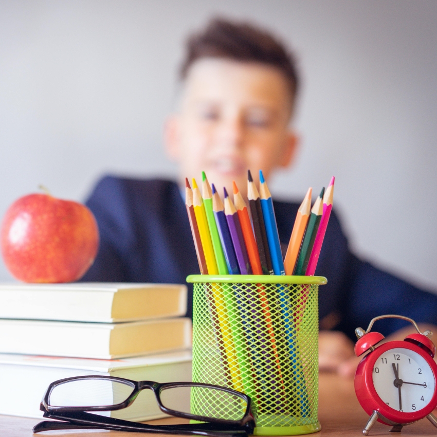 Colorful pencils in a green holder, with glasses, an apple, stacked books, and a red alarm clock nearby.